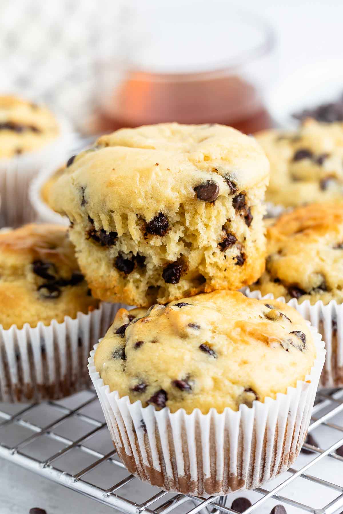 A close-up of chocolate chip muffins in white paper liners, with one muffin broken open to show the inside, all resting on a cooling rack. A glass of liquid is blurred in the background.