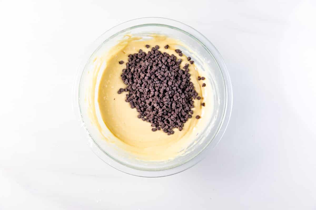 A glass bowl filled with yellow cake batter topped with a pile of mini chocolate chips sits on a white countertop.