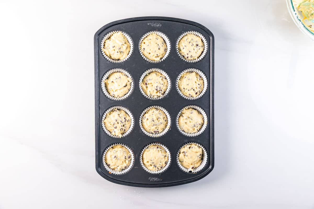 A muffin tin filled with 12 paper liners, each containing unbaked muffin batter with visible chocolate chips, sits on a white surface.
