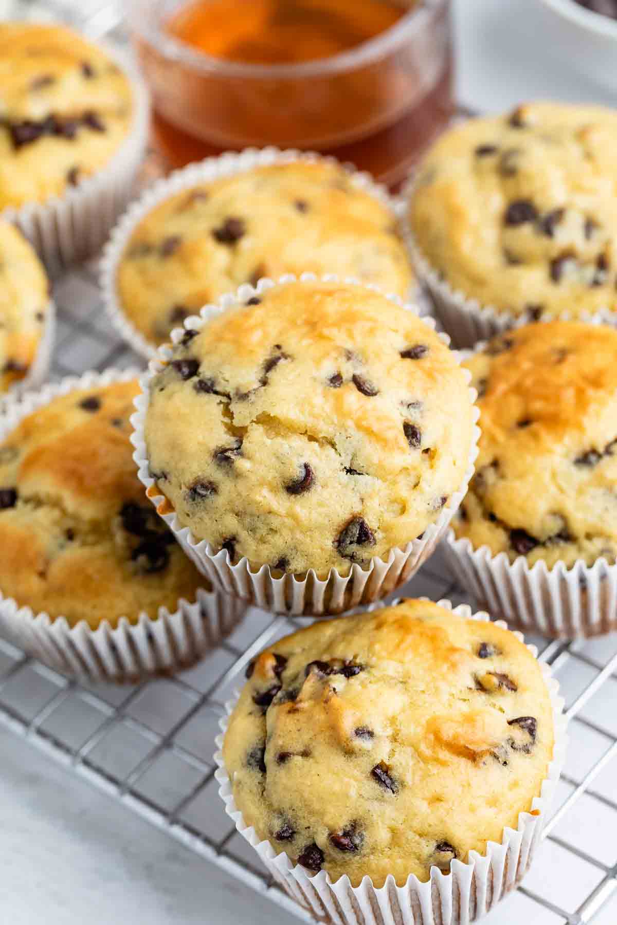 A close-up of several chocolate chip muffins in paper liners on a cooling rack, with a blurred container of honey or syrup in the background.