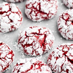 Red velvet crinkle cookies coated in powdered sugar are arranged on a light surface. The cookies have a cracked texture, showing the red interior through the white sugar coating.