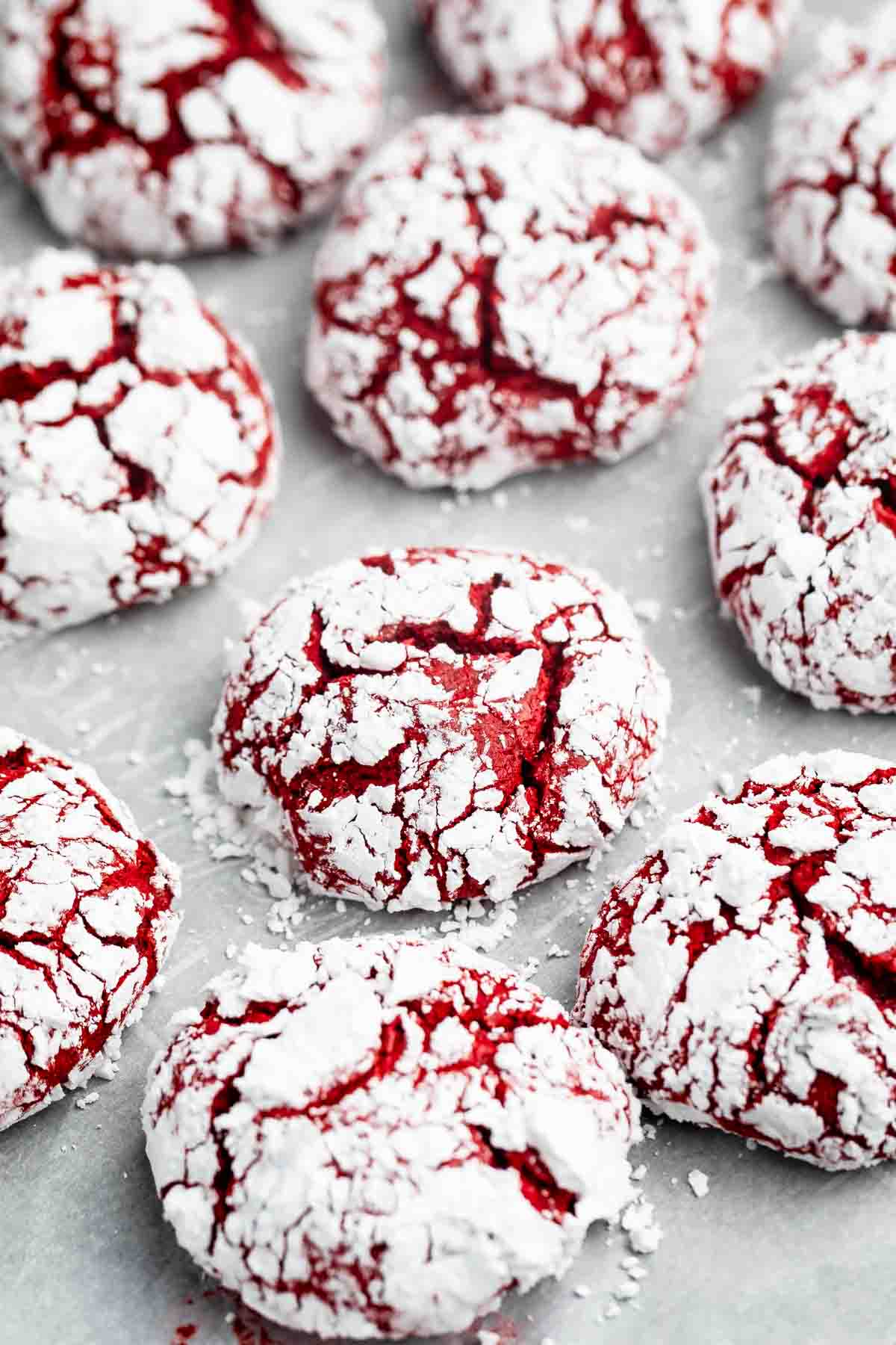 Red velvet crinkle cookies coated in powdered sugar are arranged on a light surface. The cookies have a cracked texture, showing the red interior through the white sugar coating.