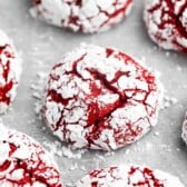 Several red velvet crinkle cookies covered in powdered sugar, with crackled surfaces revealing the red cookie beneath, displayed on a light-colored surface.