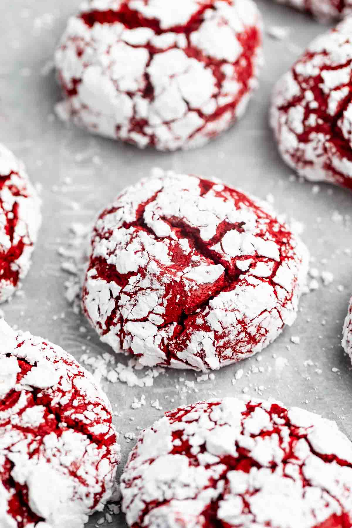 Several red velvet crinkle cookies covered in powdered sugar, with crackled surfaces revealing the red cookie beneath, displayed on a light-colored surface.