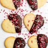 Six heart-shaped cookies partially dipped in chocolate and decorated with red, white, and pink sprinkles are arranged on a light surface. Some loose sprinkles are scattered around the cookies.