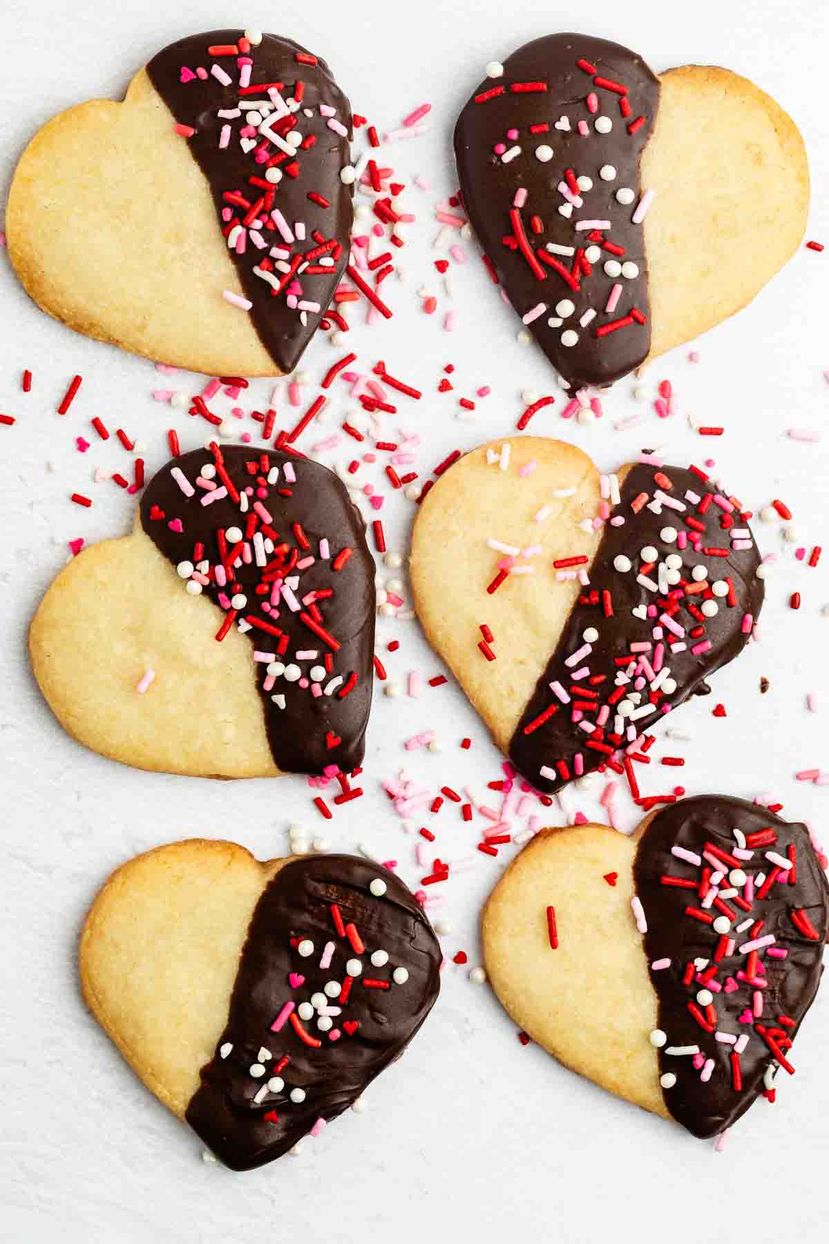 Six heart-shaped cookies partially dipped in chocolate and decorated with red, white, and pink sprinkles are arranged on a light surface. Some loose sprinkles are scattered around the cookies.
