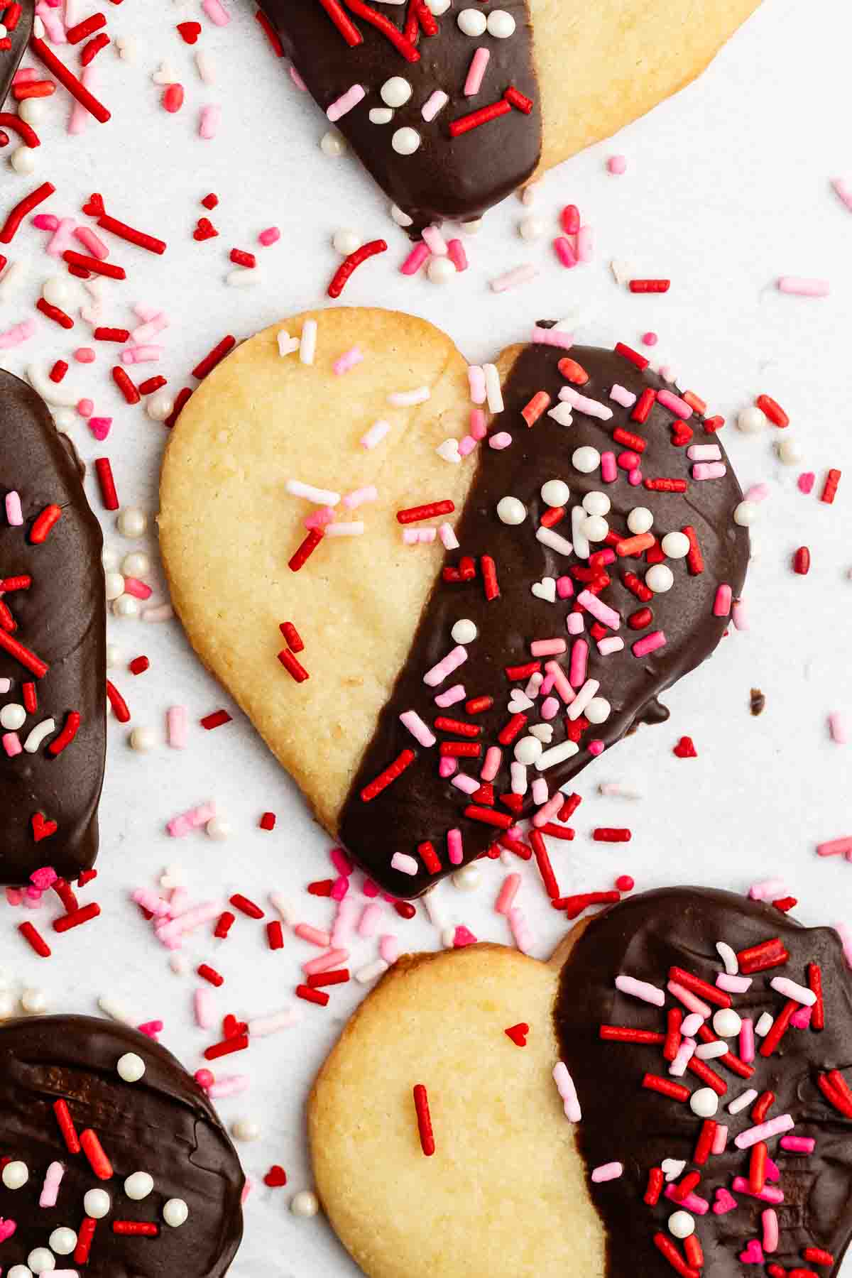 Heart-shaped cookies partially dipped in chocolate and decorated with red, pink, and white sprinkles are arranged on a white surface, with additional sprinkles scattered around them.