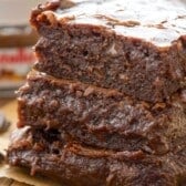 Three thick, moist slices of chocolate banana bread are stacked on parchment paper, showing a rich, fudgy texture with melted chocolate chips. A jar of Nutella is blurred in the background.