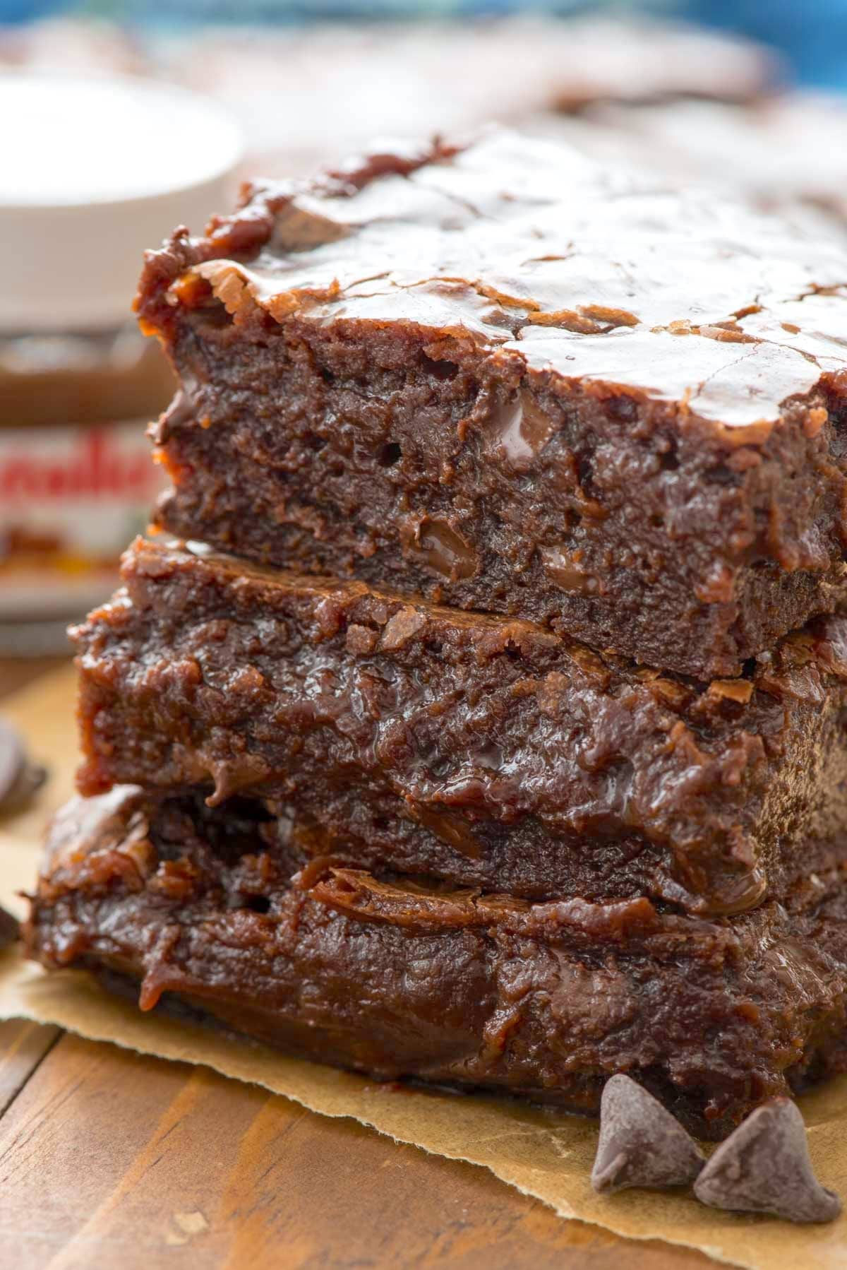 Three thick, moist slices of chocolate banana bread are stacked on parchment paper, showing a rich, fudgy texture with melted chocolate chips. A jar of Nutella is blurred in the background.
