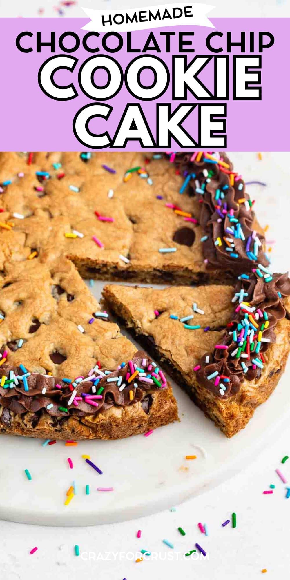 A round chocolate chip cookie cake with chocolate frosting and colorful sprinkles around the edge. A slice is cut and slightly pulled out, sitting on a white surface. Text above reads Homemade Chocolate Chip Cookie Cake.