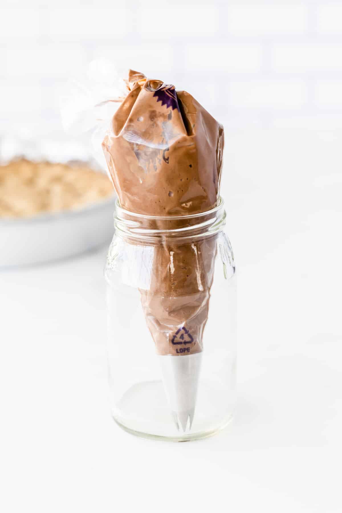 A piping bag filled with chocolate frosting is standing upright in a clear glass jar on a white surface, with a blurred pie in the background.