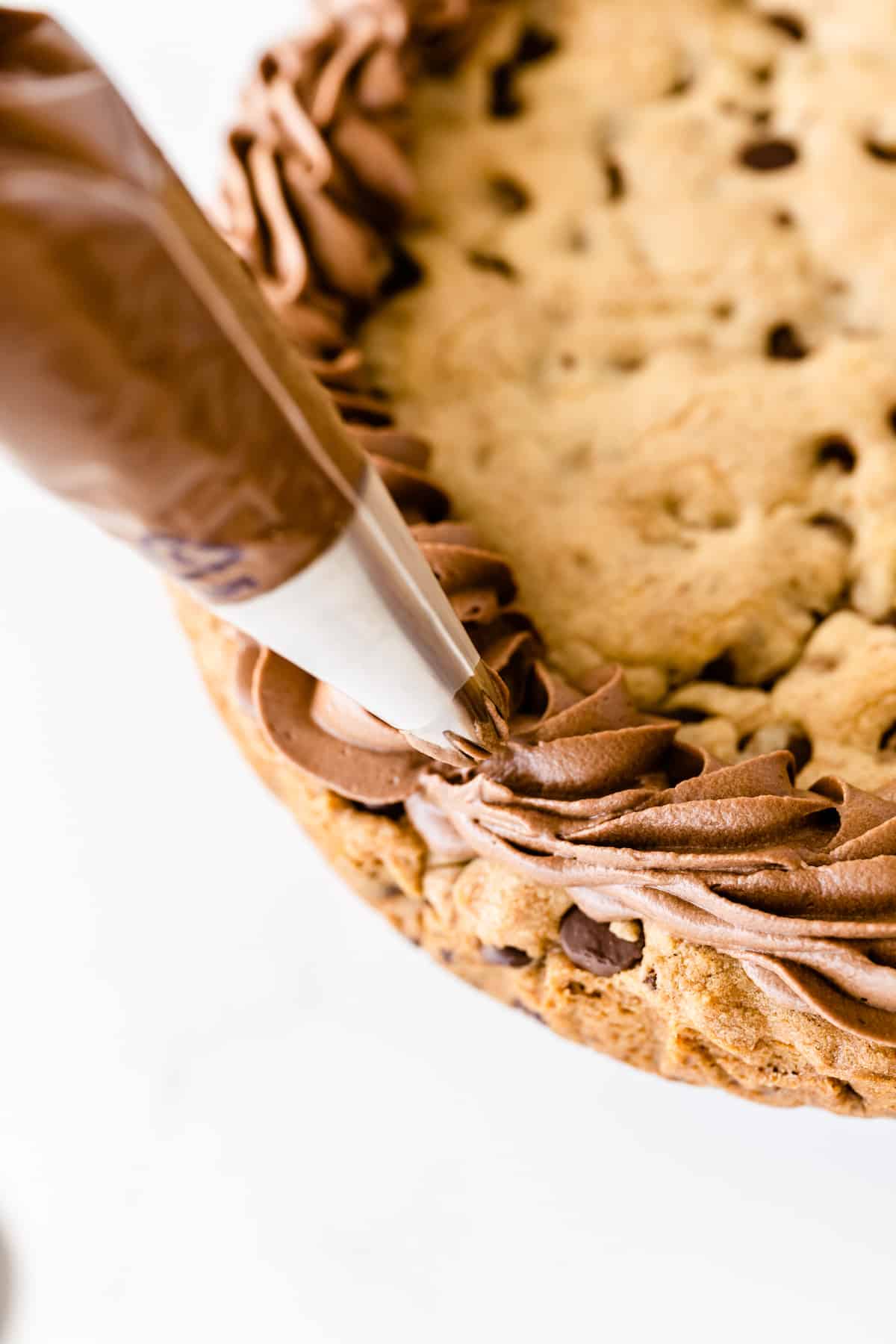 A close-up of a chocolate chip cookie cake being decorated with swirls of chocolate frosting from a piping bag along the edge.