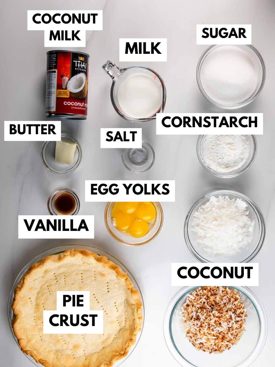 Labeled ingredients for coconut cream pie: coconut milk, milk, sugar, cornstarch, salt, butter, egg yolks, vanilla, coconut, toasted coconut, and a baked pie crust arranged on a white surface.
