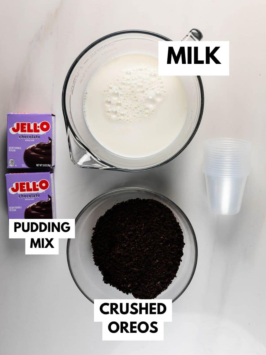 A top-down view of ingredients for a dessert: a measuring cup of milk, two boxes of Jello chocolate pudding mix, a bowl of crushed Oreos, and a stack of clear plastic cups on a white surface. Labels identify each item.