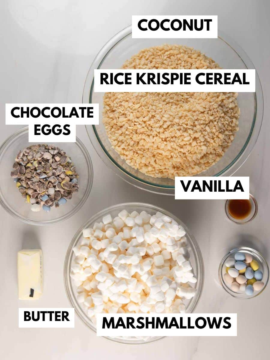 Five labeled bowls and ingredients on a white surface: shredded coconut, rice krispie cereal, chopped chocolate eggs, vanilla extract, a stick of butter, and mini marshmallows.