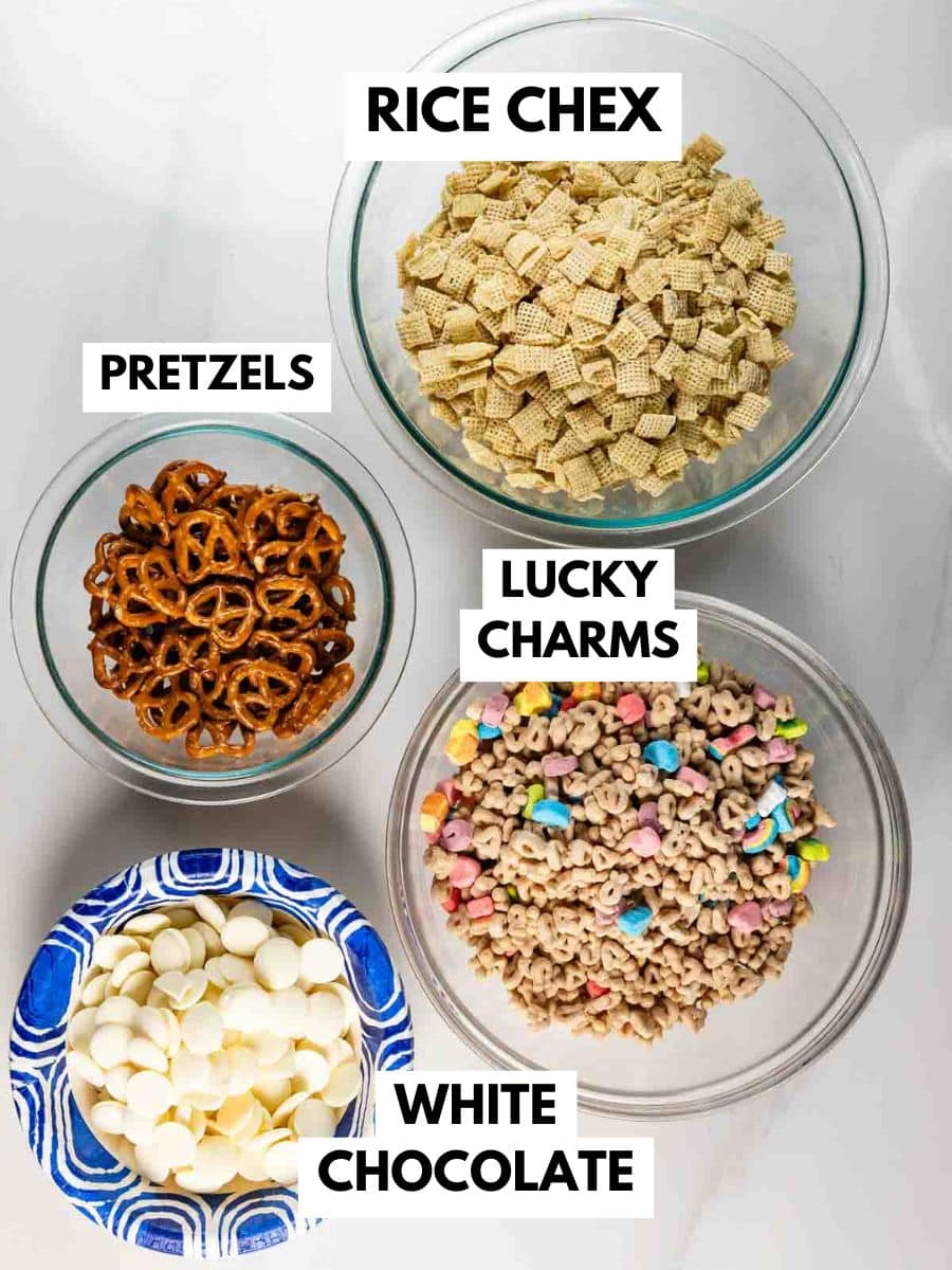 Four bowls of snacks labeled: Rice Chex cereal, pretzels, Lucky Charms cereal, and white chocolate chips, arranged on a white surface.