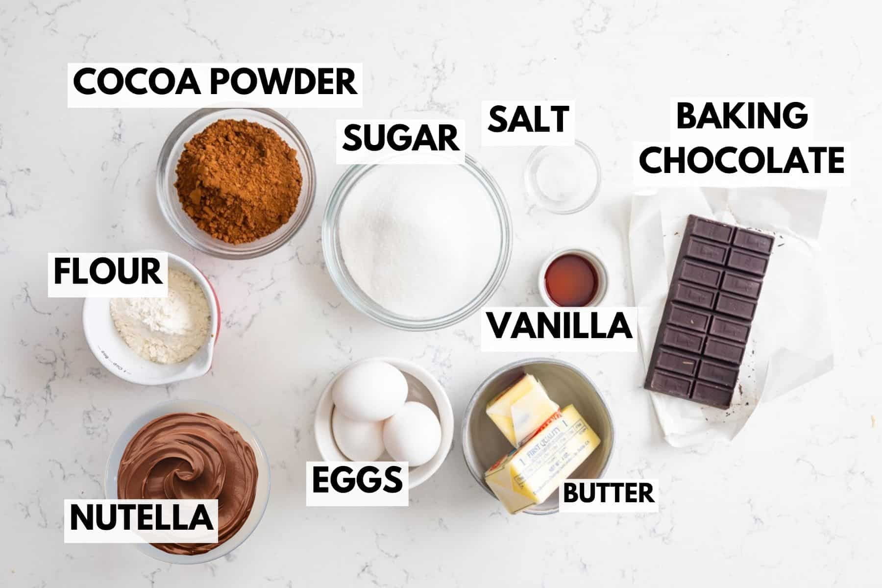 Top view of labeled baking ingredients on a white surface, including bowls of cocoa powder, flour, sugar, salt, Nutella, eggs, butter, vanilla extract, and a bar of baking chocolate.