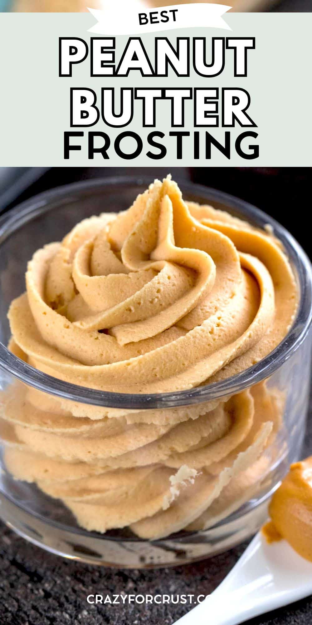 A swirl of creamy peanut butter frosting in a clear glass cup, with a white spoon holding a dollop of frosting beside it. The text above reads Best Peanut Butter Frosting.