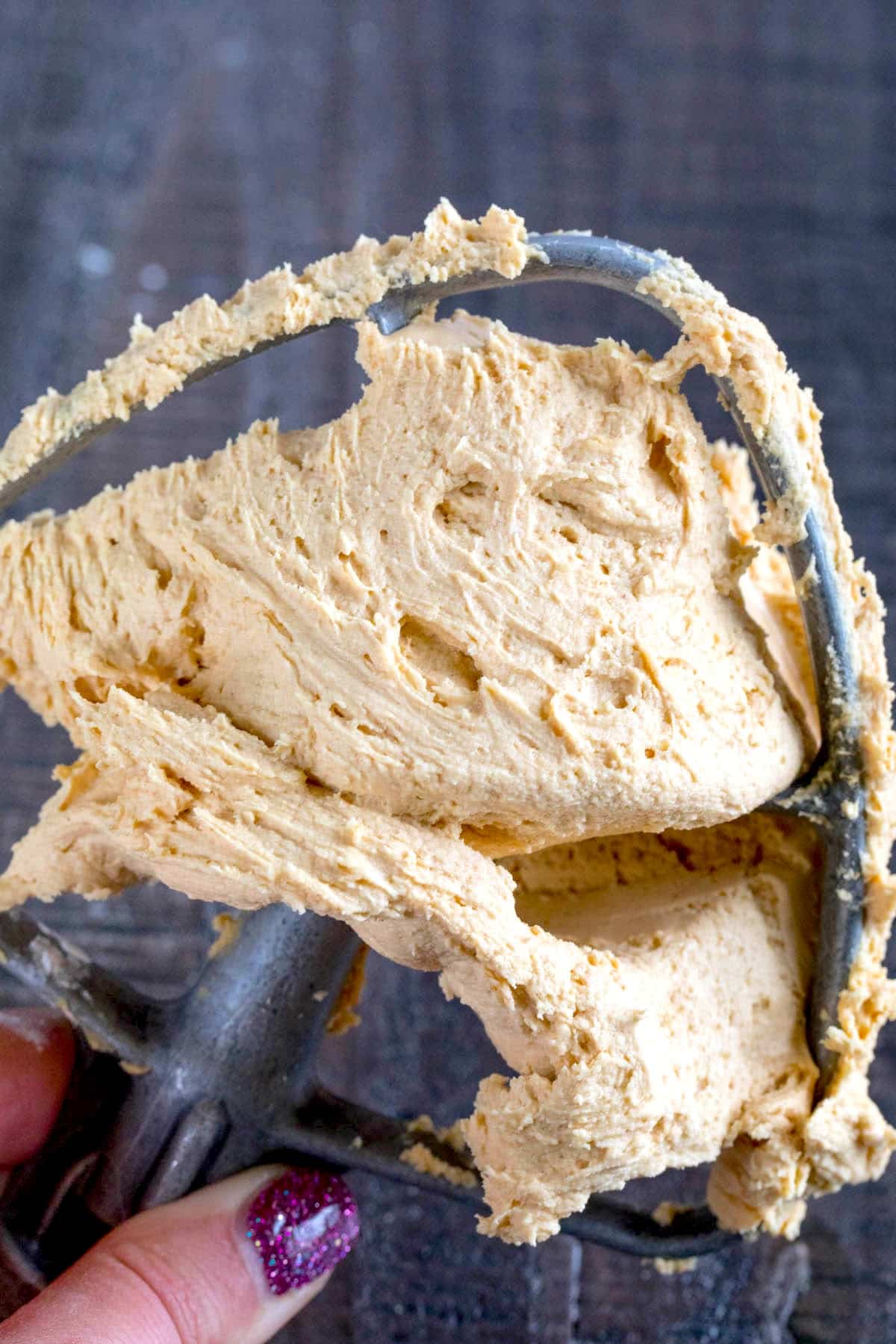 A hand holds a mixer beater covered in creamy, light brown frosting against a dark background. The person’s fingernails are painted with sparkly purple nail polish.