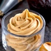 A clear glass cup filled with swirled peanut butter frosting, with a piping bag and chocolate cupcake in the background, and a spoonful of peanut butter nearby.
