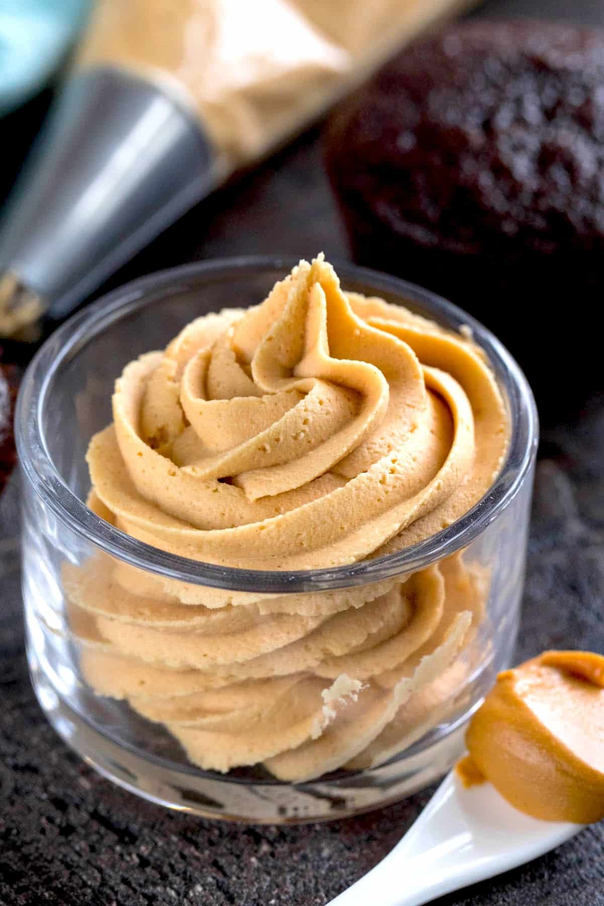 A clear glass cup filled with swirled peanut butter frosting, with a piping bag and chocolate cupcake in the background, and a spoonful of peanut butter nearby.