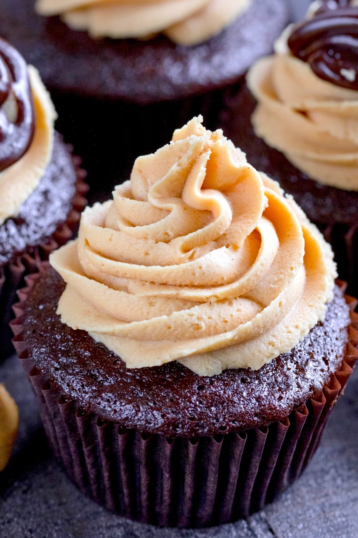 A close-up of a chocolate cupcake with a swirl of creamy light brown frosting on top, set in a dark brown paper liner. Other similar cupcakes are visible in the background.
