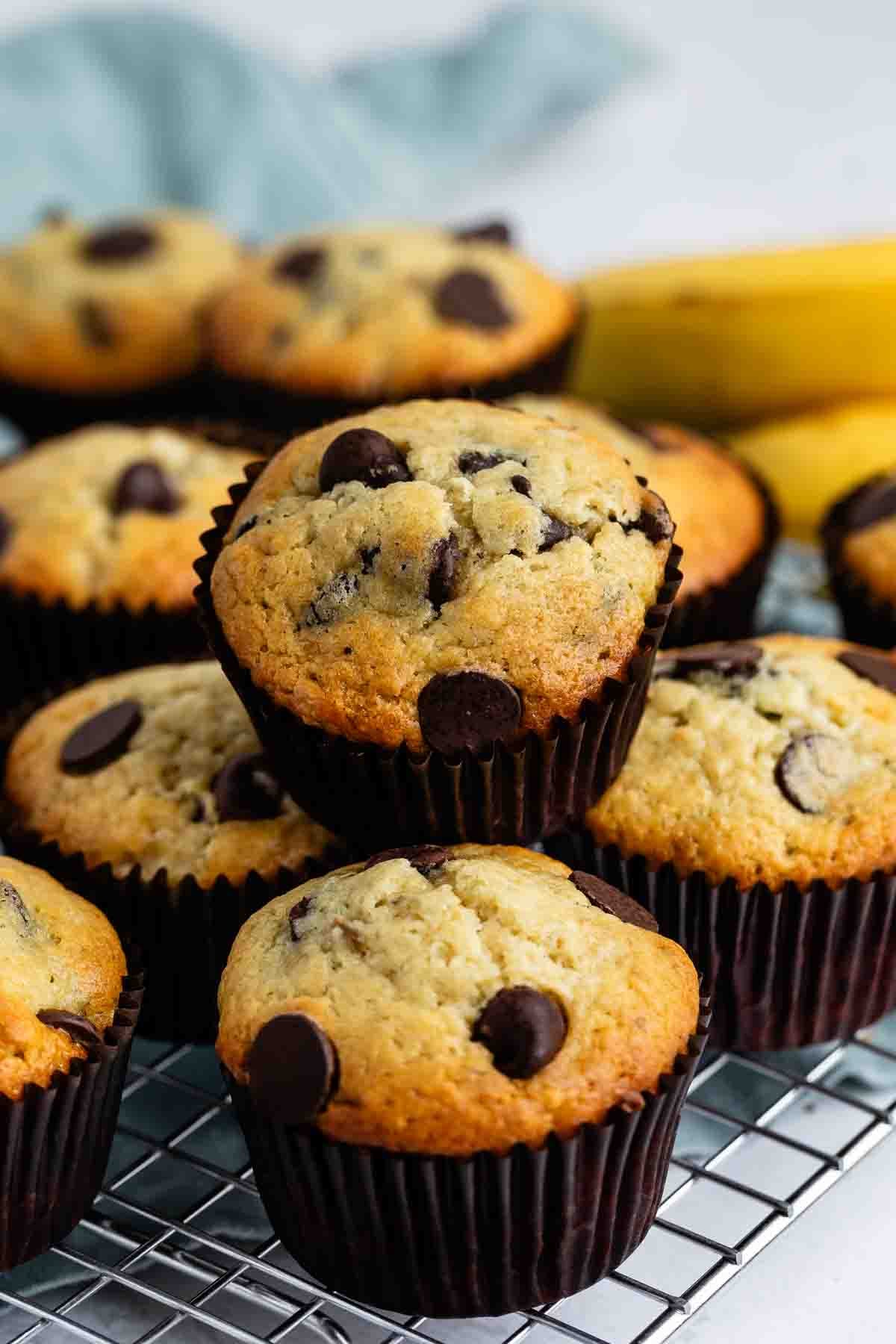 A close-up of several freshly baked banana chocolate chip muffins in dark liners, stacked on a wire cooling rack with a blurred banana and cloth in the background.