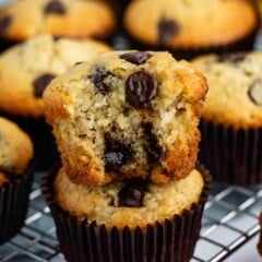 A stack of two chocolate chip muffins on a cooling rack, with the top muffin broken in half showing a moist interior filled with chocolate chips. More muffins are visible in the background.