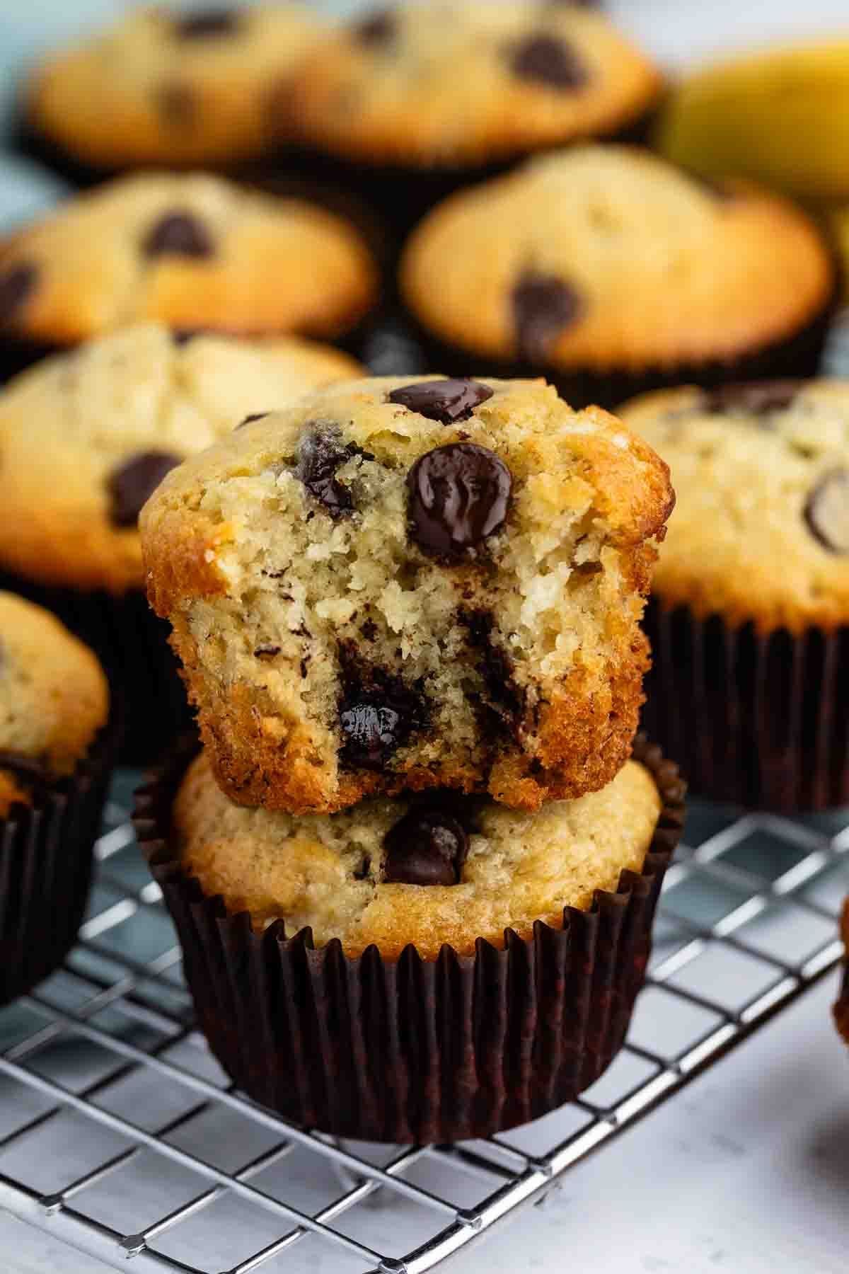 A stack of two chocolate chip muffins on a cooling rack, with the top muffin broken in half showing a moist interior filled with chocolate chips. More muffins are visible in the background.