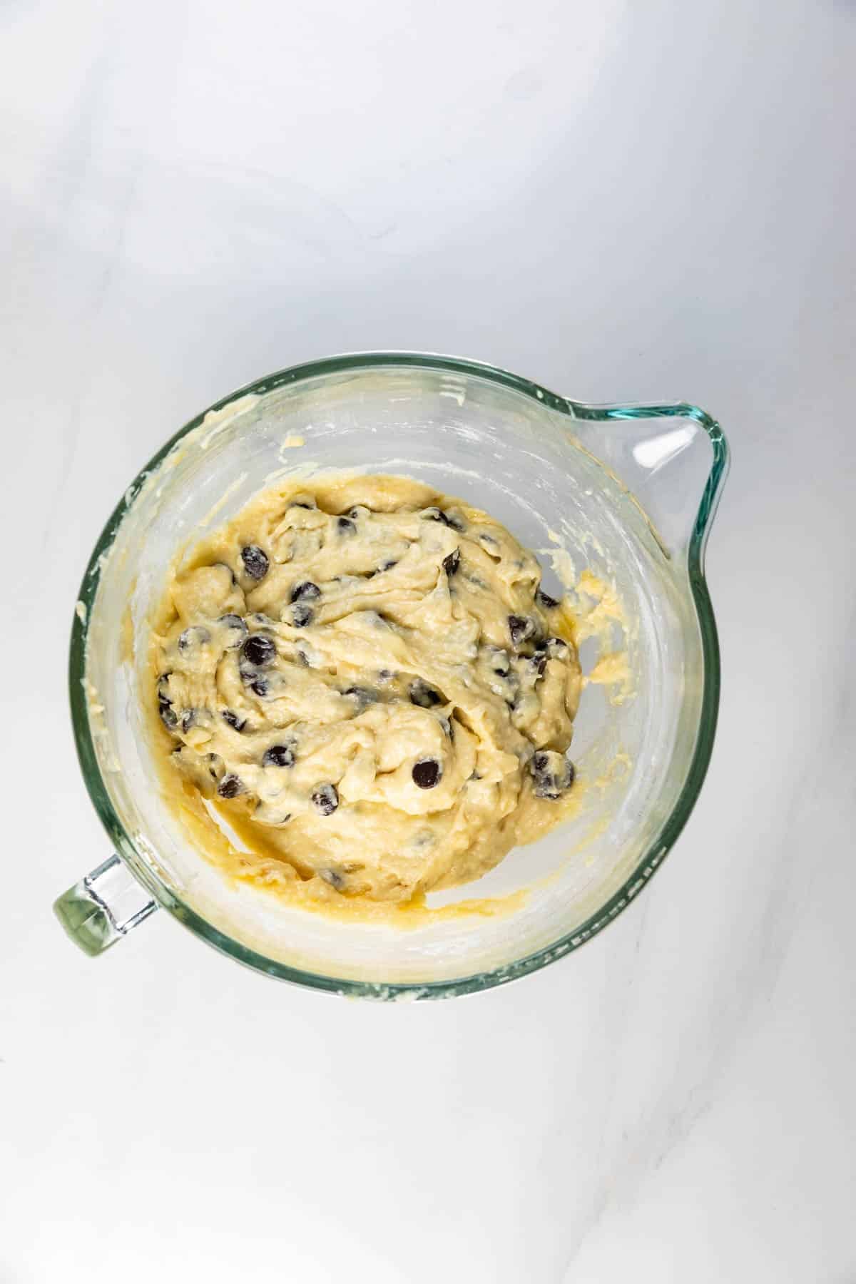 A glass mixing bowl filled with chocolate chip cookie dough sits on a white marble surface.