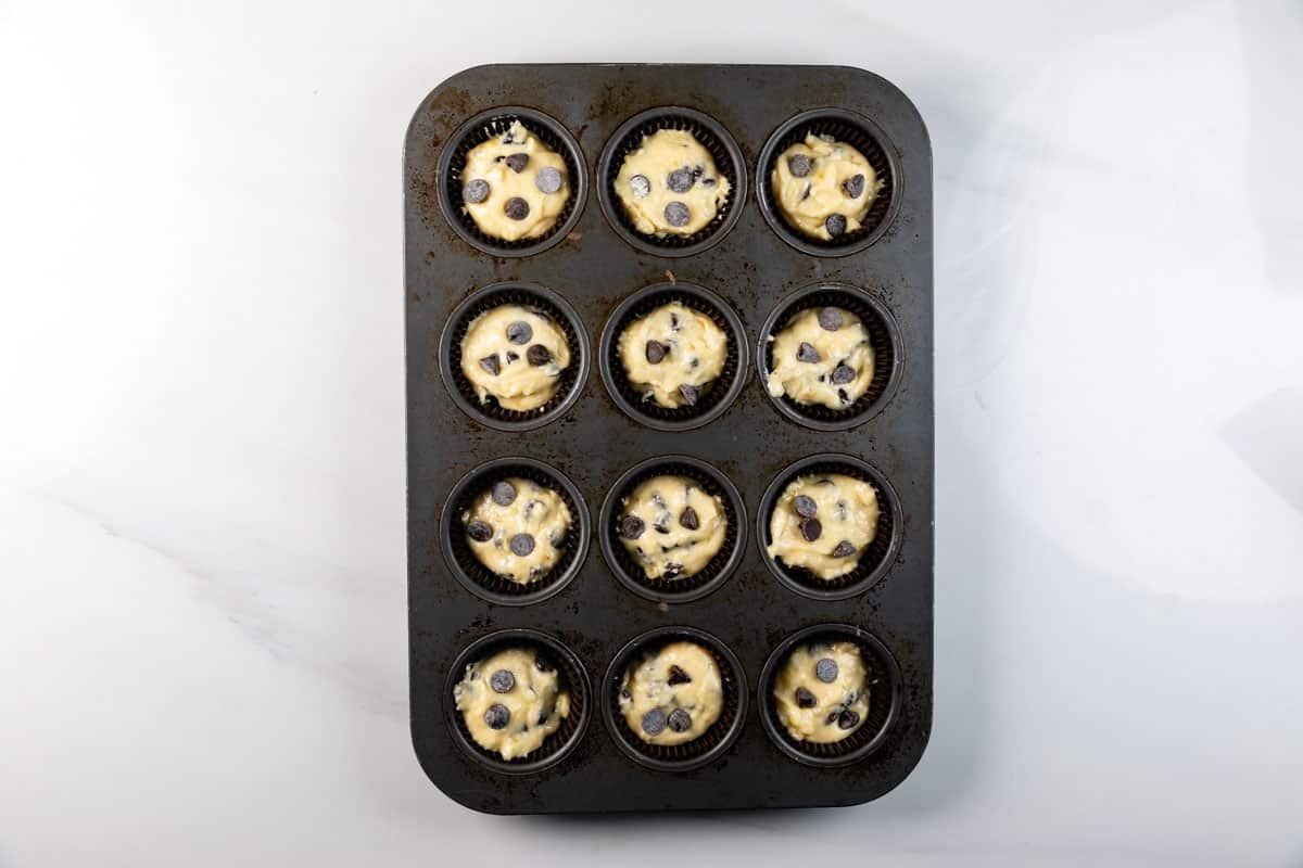 A muffin tin with twelve cups, each filled with unbaked muffin batter and topped with chocolate chips, sits on a white surface.