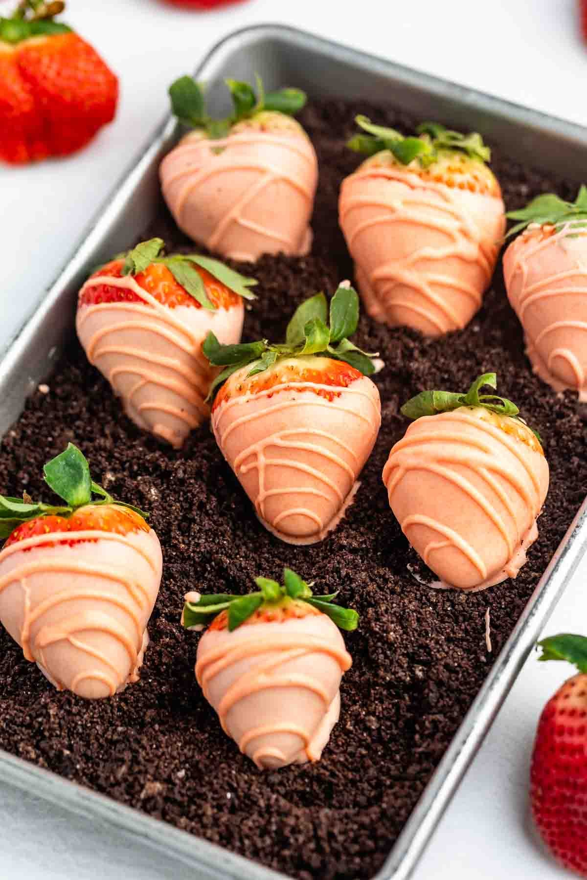 A tray of strawberries dipped in peach-colored chocolate and drizzled with more chocolate, arranged on a bed of crumbled chocolate cookies. The strawberries have green tops and are neatly lined up.