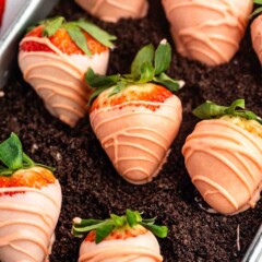 Strawberries dipped in light pink chocolate with a drizzle design, arranged on a bed of crumbled chocolate cookie crumbs in a metal tray.