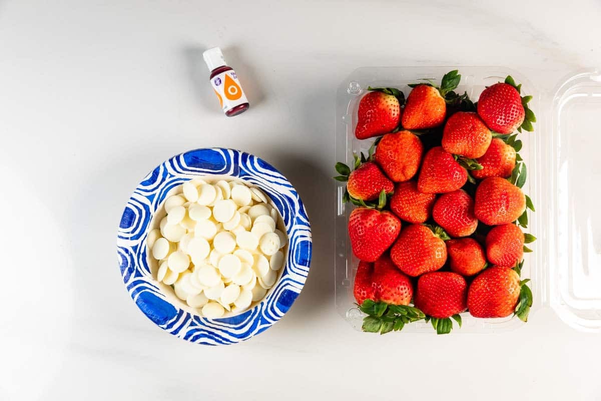 A carton of fresh strawberries, a bowl of white chocolate melting wafers, and a small bottle of orange oil are arranged on a white surface.