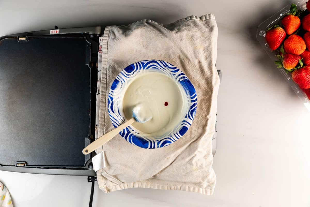 A bowl of white chocolate with a single drop of red food coloring, a spatula inside, placed on a cloth next to a closed griddle and a carton of fresh strawberries on a white surface.