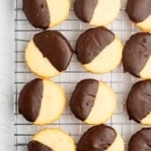 Round shortbread cookies half-dipped in chocolate are arranged neatly on a cooling rack, displayed on a light-colored surface.