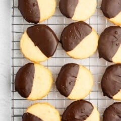 Round shortbread cookies half-dipped in chocolate are arranged neatly on a cooling rack, displayed on a light-colored surface.