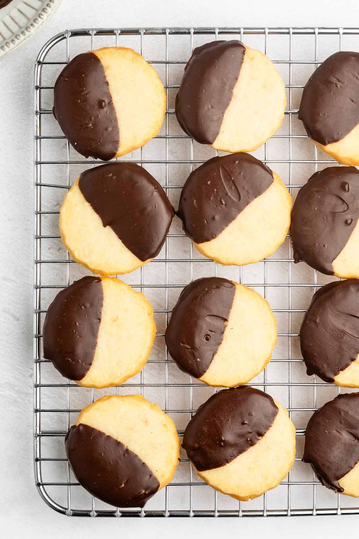Round shortbread cookies half-dipped in chocolate are arranged neatly on a cooling rack, displayed on a light-colored surface.