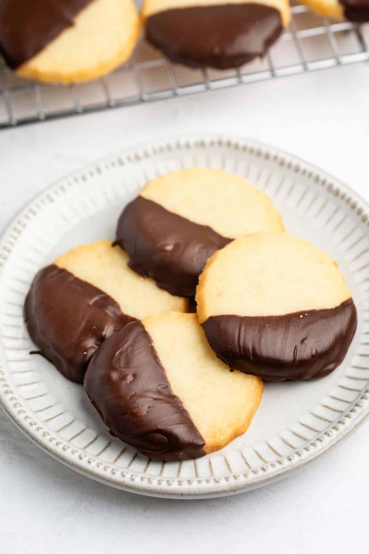A plate with four round shortbread cookies, each half-dipped in chocolate, sitting on a white plate with a textured rim. More cookies are visible on a cooling rack in the background.