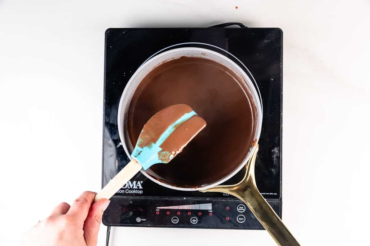 A hand holding a blue spatula stirs thick melted chocolate in a saucepan on an electric stovetop viewed from above.