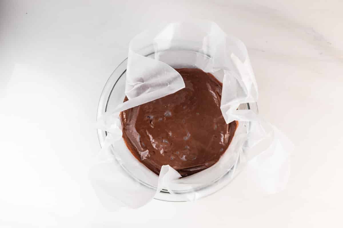 A glass bowl lined with parchment paper is filled with chocolate cake batter, sitting on a white countertop.