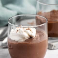 A clear glass filled with chocolate pudding, topped with a dollop of whipped cream and chocolate shavings, sits on a white surface with another pudding glass and a light blue cloth in the background.