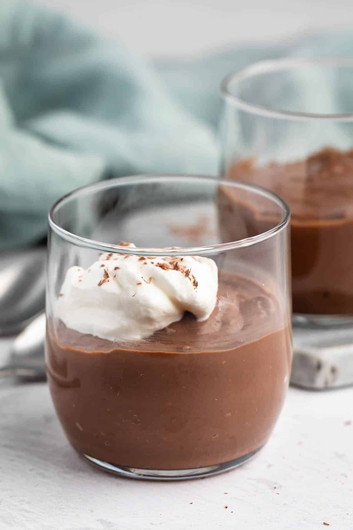 A clear glass filled with chocolate pudding, topped with a dollop of whipped cream and chocolate shavings, sits on a white surface with another pudding glass and a light blue cloth in the background.