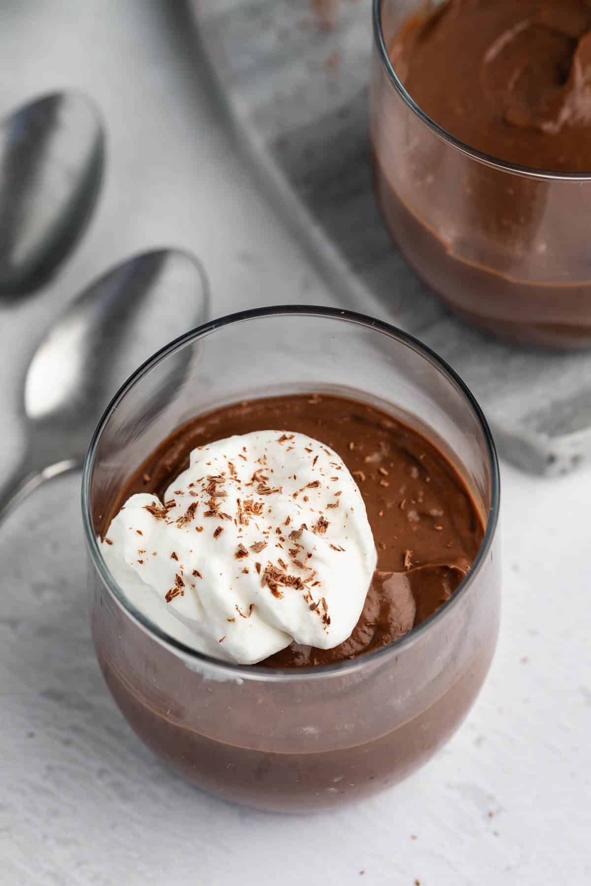 A glass cup filled with chocolate pudding, topped with a dollop of whipped cream and chocolate shavings, sits on a white surface next to two metal spoons and another cup in the background.