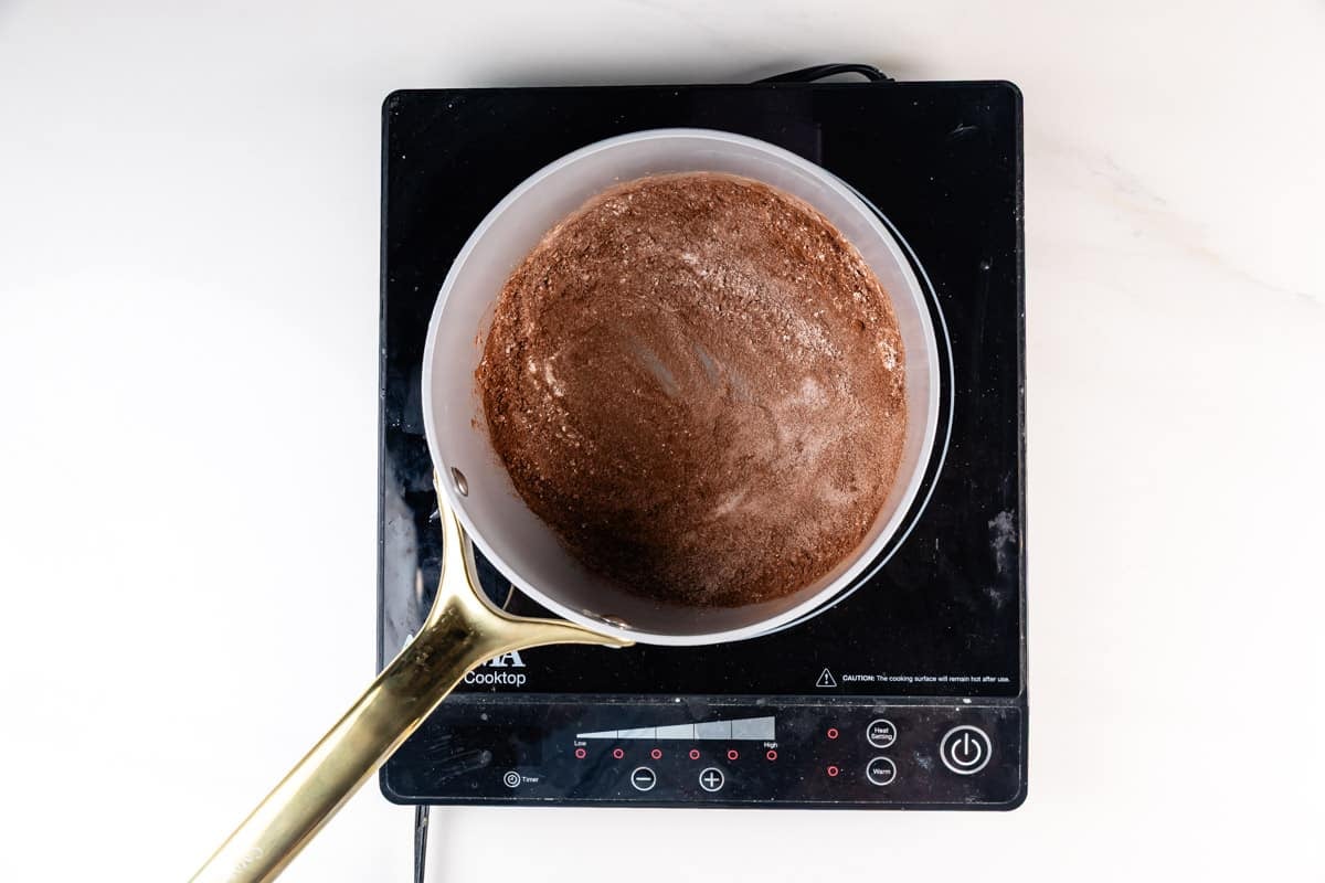 A saucepan with a golden handle filled with a brown cocoa mixture is placed on a black induction cooktop against a white background.