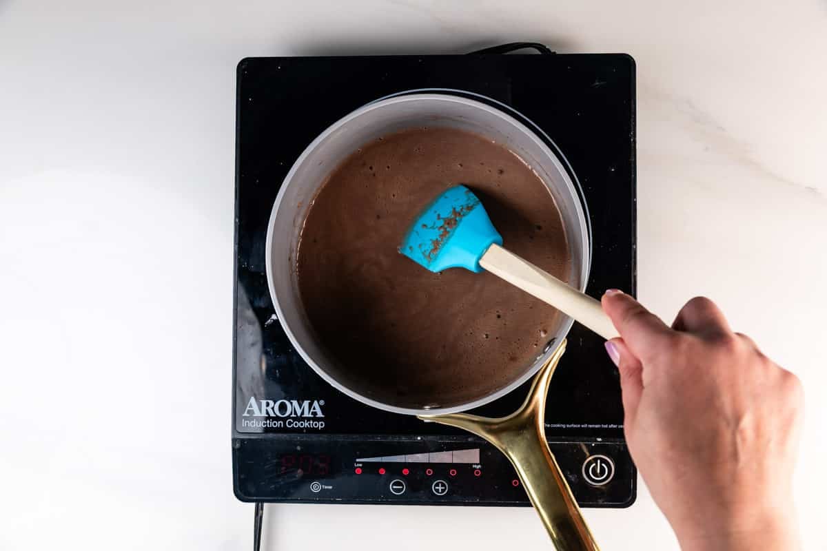 A person stirs chocolate mixture in a saucepan with a blue spatula on an Aroma induction cooktop.