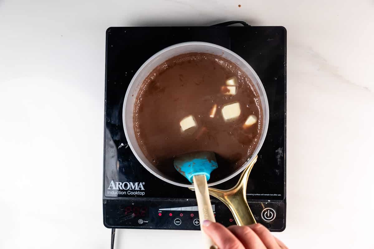 A hand stirs a pot of chocolate mixture with melting butter chunks on an induction cooktop. The cooktop display shows 00:0 and the spatula being used is blue.