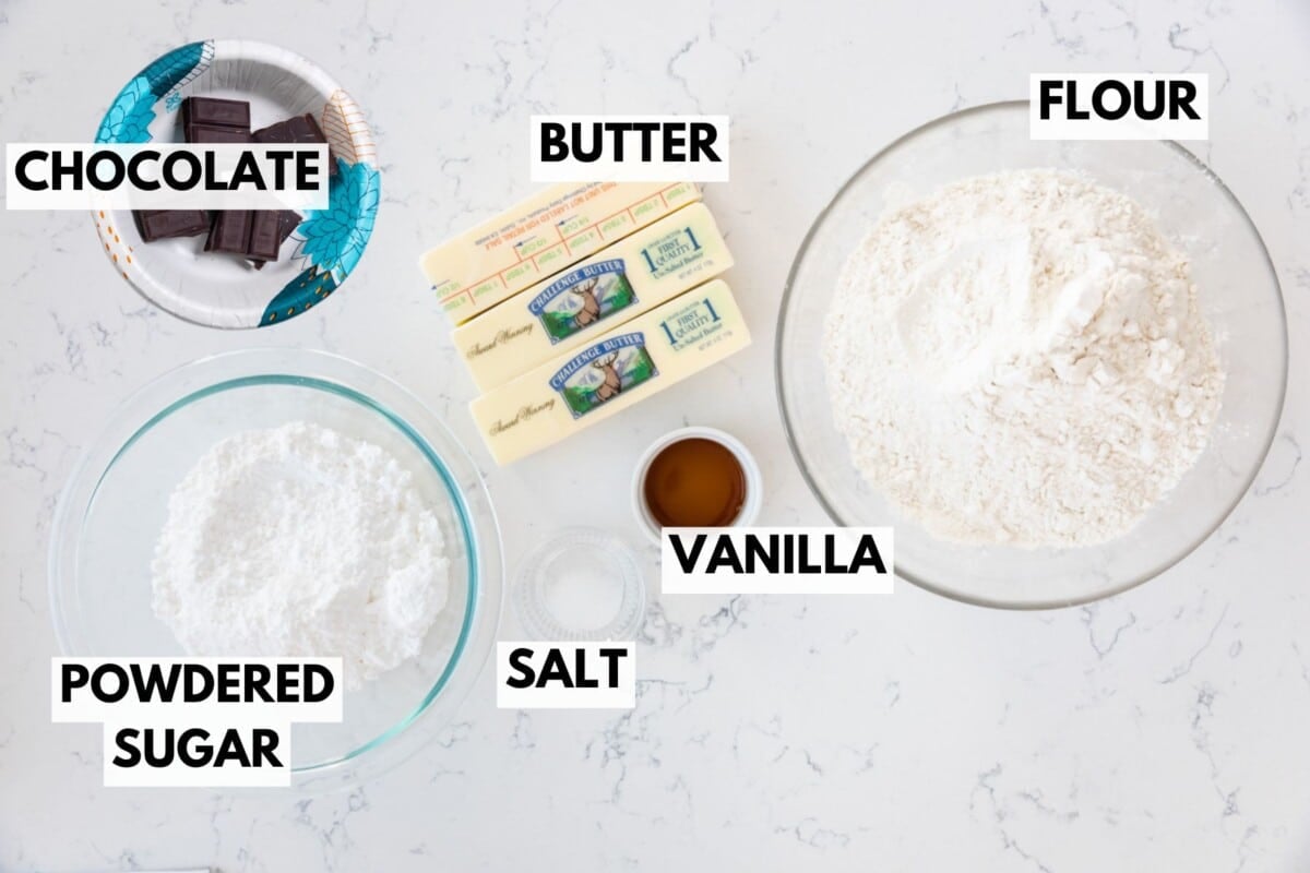 Overhead view of baking ingredients on a white surface: a plate of chocolate squares, sticks of butter, a bowl of flour, a bowl of powdered sugar, a small bowl of vanilla extract, and a pinch of salt. Each is labeled.