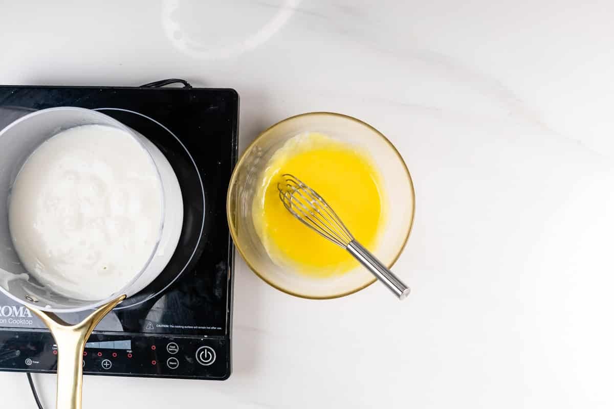 A bowl of melted butter with a whisk next to a saucepan of milk heating on an electric cooktop, all on a white countertop.