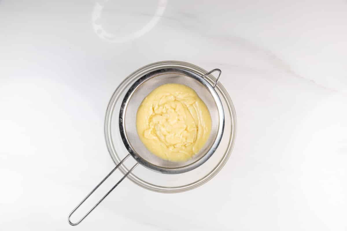 A metal strainer holding thick yellow custard sits on top of a glass bowl, all placed on a white marble surface.